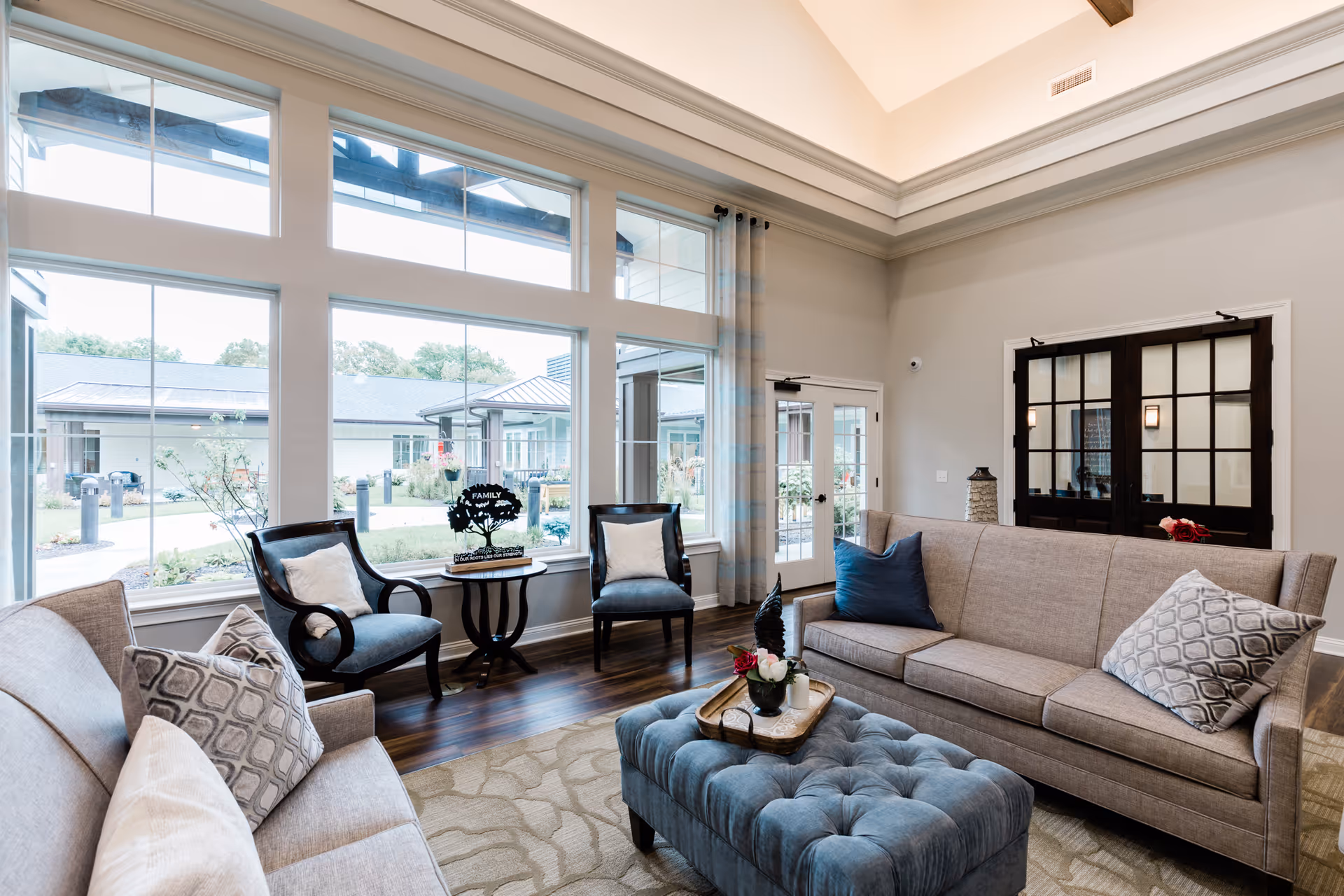 A bright and spacious living room with large windows overlooking an outdoor courtyard. The room features two beige sofas with patterned cushions, two dark wooden chairs with light cushions, a blue tufted ottoman with a tray holding flowers, and a small round table with a decorative family tree sign. The walls are light-colored, and there are double glass doors with dark frames on one side.