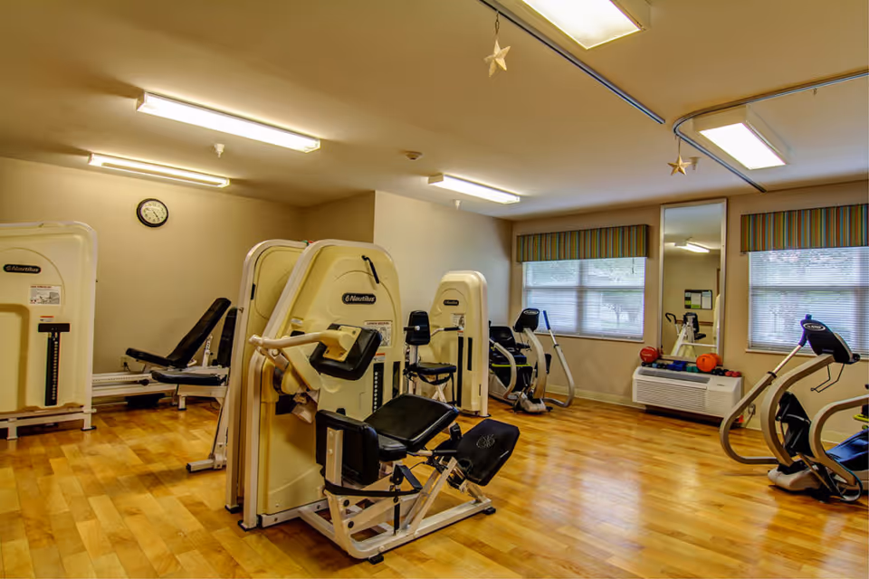 A fitness room with various exercise machines including stationary bikes and strength training equipment. The room has wooden flooring, beige walls, fluorescent ceiling lights, two windows with striped valances, and a large mirror on one wall. There are some small exercise balls and weights on a shelf beneath the windows.