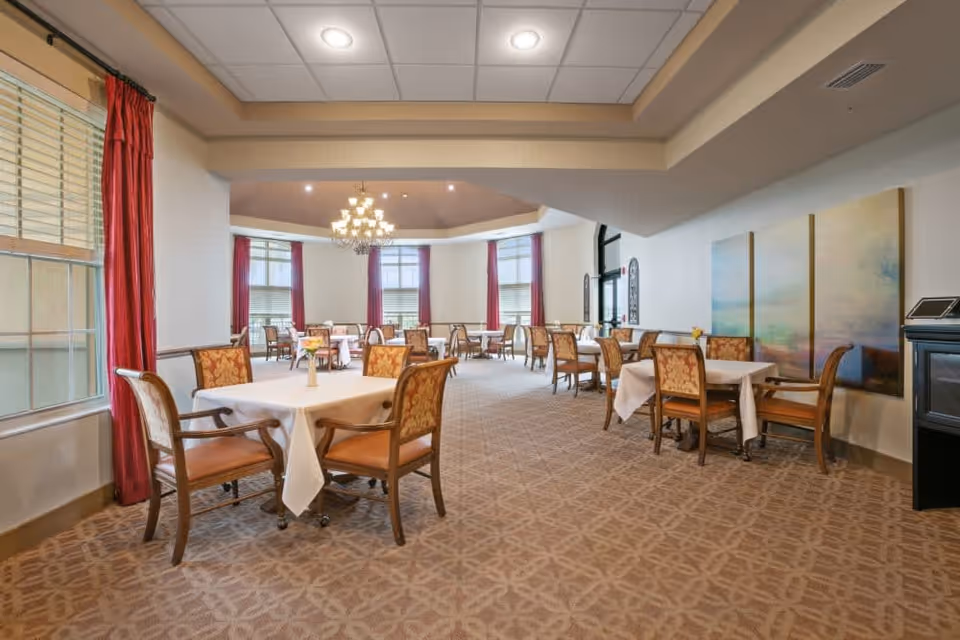 A spacious dining room with multiple tables covered in white tablecloths, each surrounded by four upholstered chairs. The room features large windows with red curtains, a patterned carpet, and a chandelier hanging from the ceiling. There are three large abstract paintings on one wall and a small cabinet with a tablet device on top.