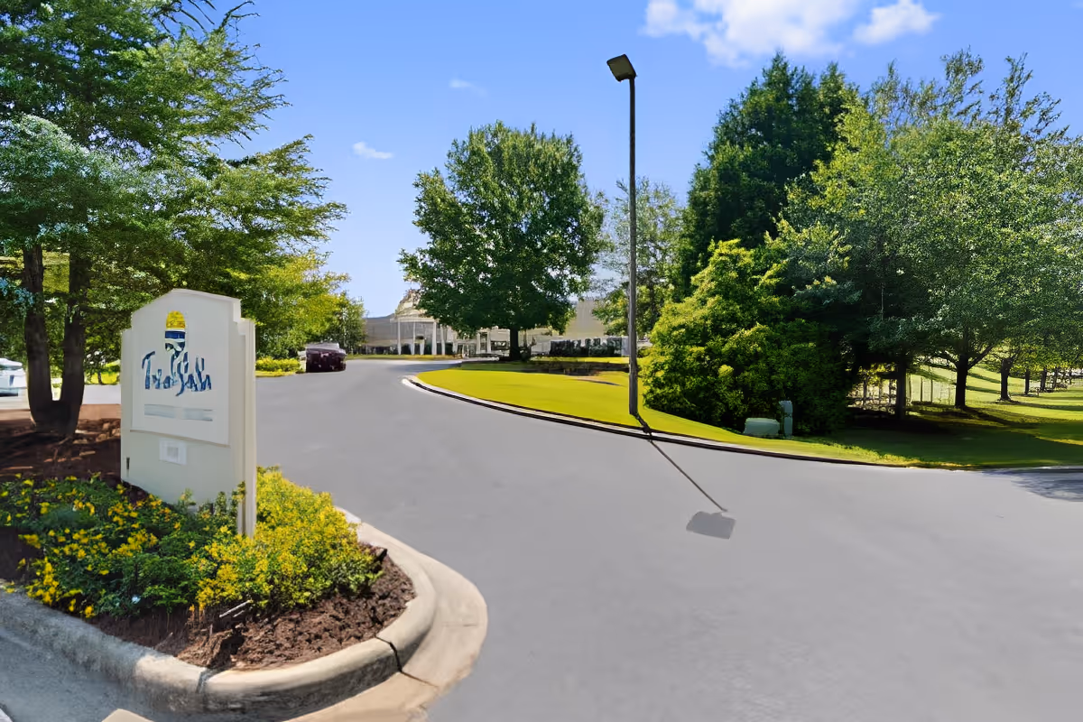 Curved driveway and landscaped entrance with a sign, lamp post, and trees leading to a building in the background.