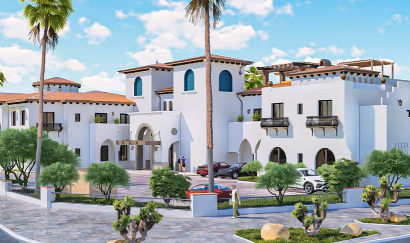 Exterior view of a large, white Mediterranean-style building with arched windows and doorways, surrounded by palm trees, shrubs, and a paved driveway with parked cars. The sky is blue with scattered clouds.