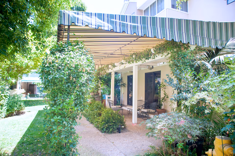 Outdoor patio area at The Heritage Downtown with a striped awning, surrounded by lush green plants and trees. There are chairs and small tables under the awning next to the building.