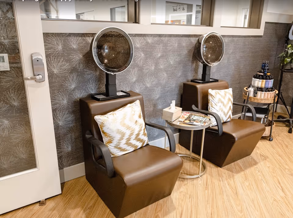 Two brown salon chairs with hooded hair dryers, patterned pillows, and a small side table in a salon seating area.