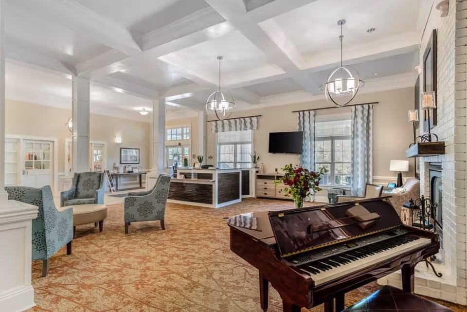 Bright communal living room with a grand piano, seating area, and a reception desk under a coffered ceiling.