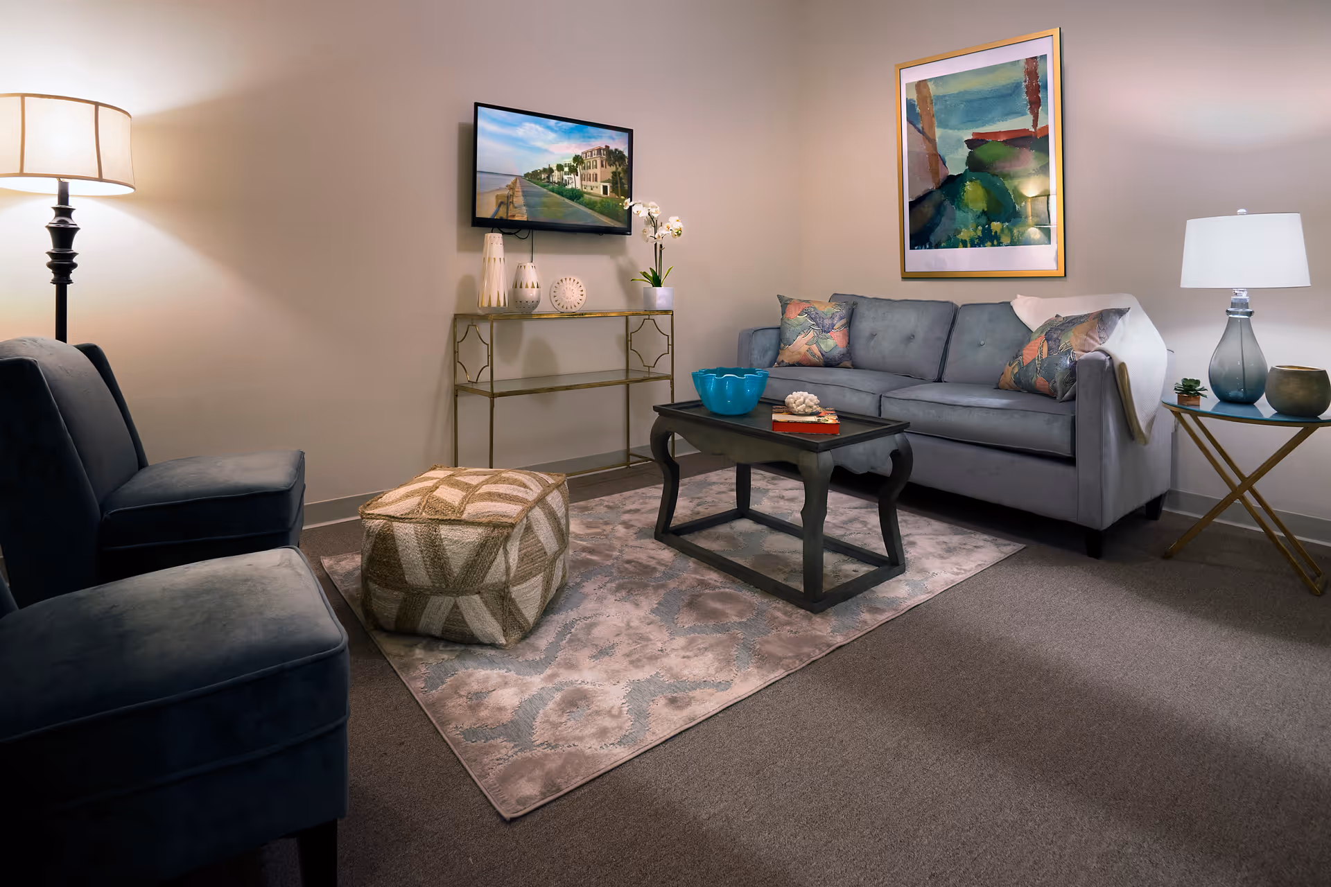 A cozy living room with a gray sofa adorned with patterned pillows, a dark wooden coffee table with decorative items, a patterned pouf, two blue upholstered chairs, a floor lamp, a side table with a lamp and decor, a wall-mounted TV, and framed artwork on the walls.