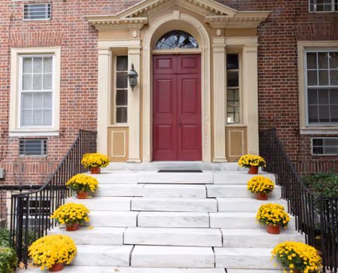 White marble steps lined with potted yellow flowers lead up to a red entrance door in a brick building.