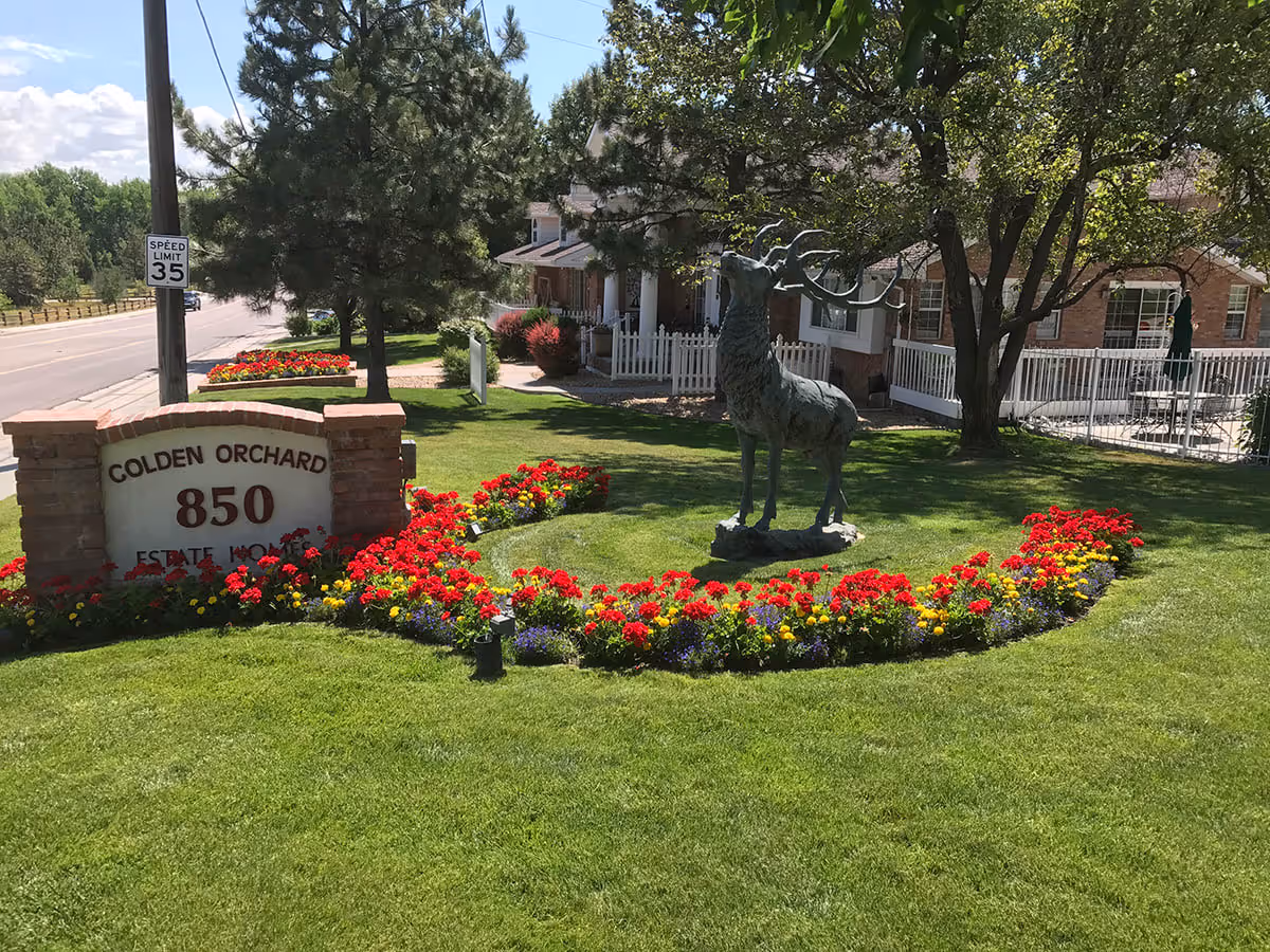 Outdoor view of Golden Orchard Estate Homes with a brick sign displaying the address 850, surrounded by a well-maintained lawn and colorful flower beds. A large metal statue of a deer stands on the grass near the sign, with trees and a building with a white fence in the background.
