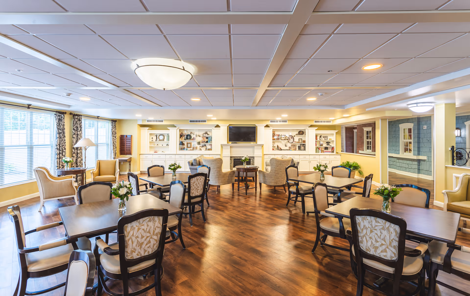 Bright communal dining and lounge area with several tables and chairs, armchairs by windows, and a fireplace with a TV on the far wall.