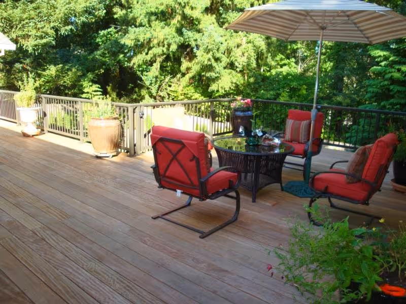 Outdoor wooden deck with four red cushioned chairs arranged around a round glass-top table. A large striped umbrella provides shade, and several potted plants are placed along the railing. Lush green trees and foliage surround the deck.