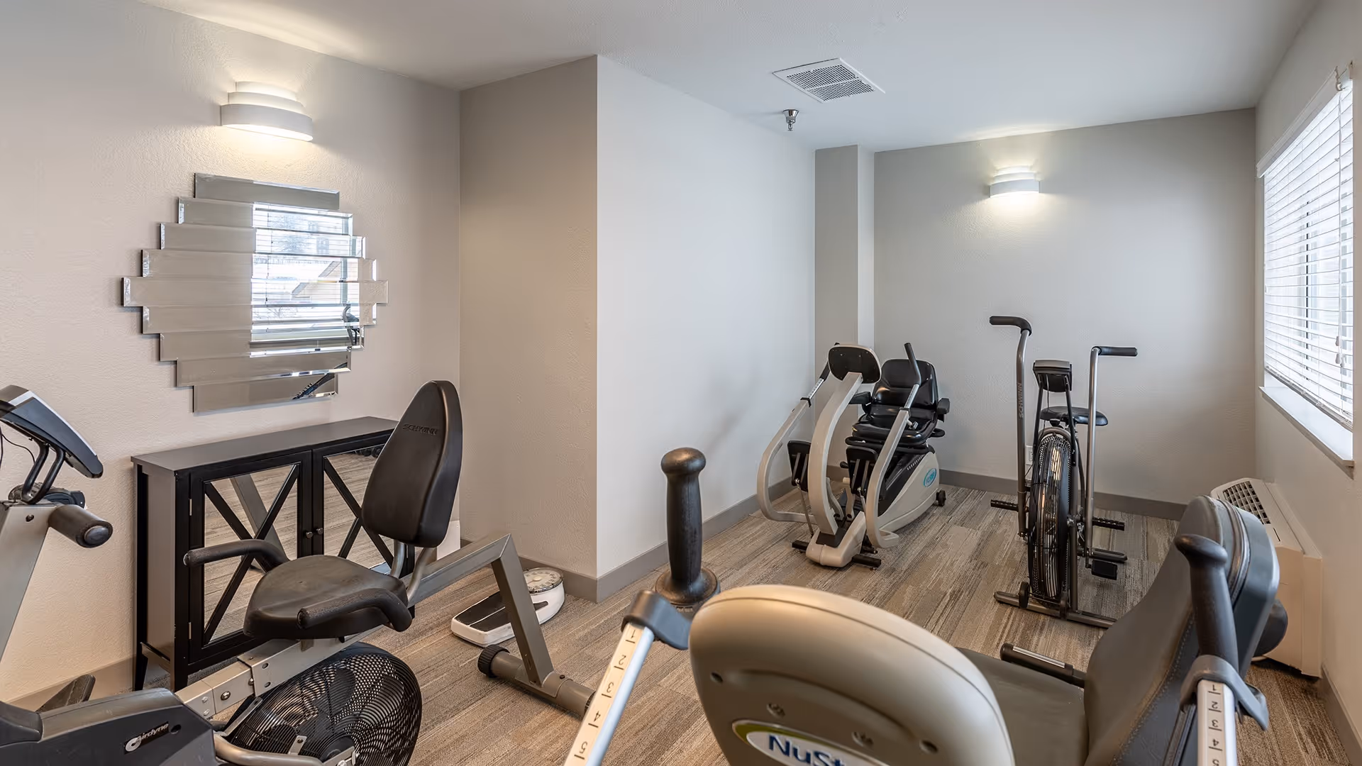 A small exercise room with several stationary exercise bikes and a recumbent bike. The room has light-colored walls, a window with blinds on the right side, a decorative mirror on the left wall above a small cabinet, and two wall-mounted lights providing illumination.