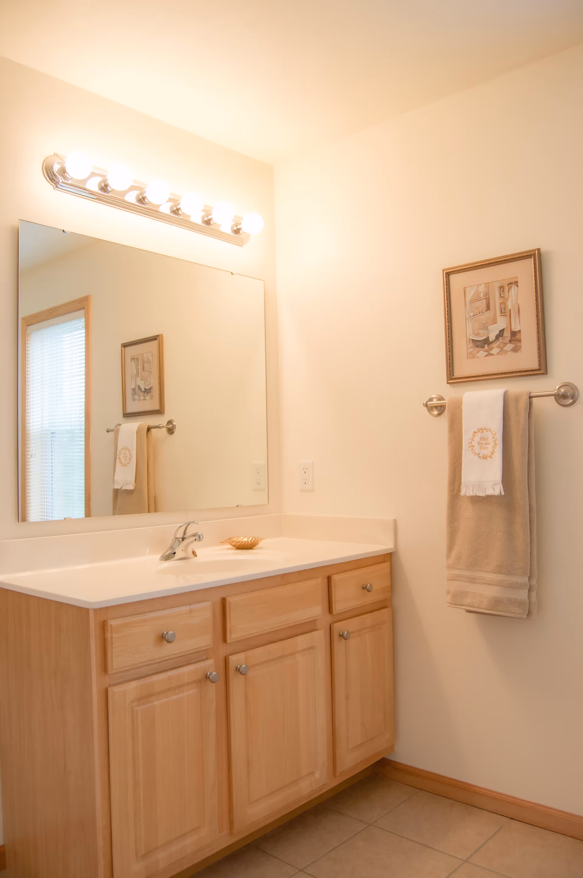 A bathroom vanity with a large mirror above it, six light bulbs mounted on a fixture above the mirror, a single faucet on the sink, and wooden cabinets below. A beige towel and a white hand towel hang on a towel rack on the wall next to the vanity, with a framed picture above the towels. The walls are light-colored and the floor is tiled.