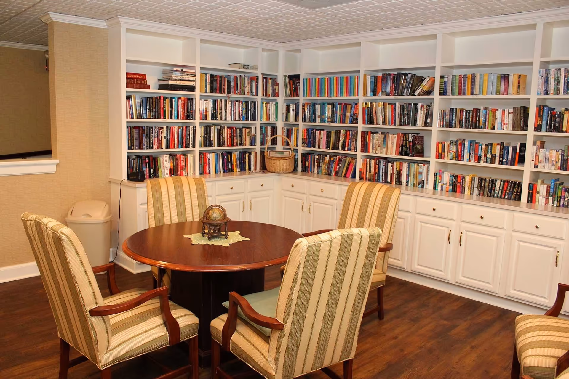 A cozy reading room with a round wooden table surrounded by four striped upholstered chairs. Behind the table is a large white built-in bookshelf filled with numerous books. The room has wooden flooring and beige walls, creating a warm and inviting atmosphere.