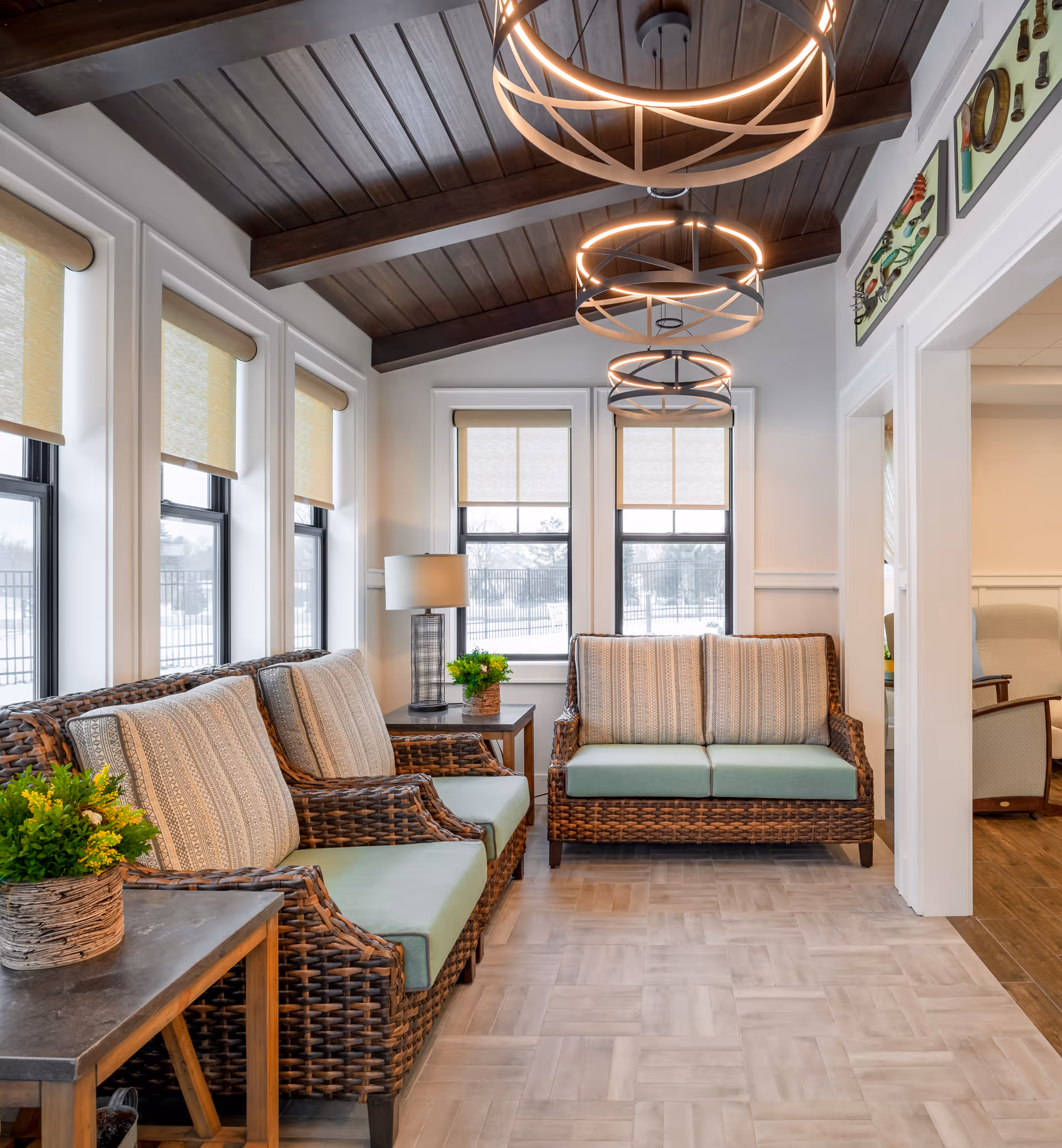 A bright and cozy sitting area with wicker sofas featuring light green cushions and patterned pillows. The room has large windows with beige roller shades, a wooden ceiling with exposed beams, and modern circular pendant lights. There are side tables with potted plants and a lamp, and the floor is covered with light-colored tiles.