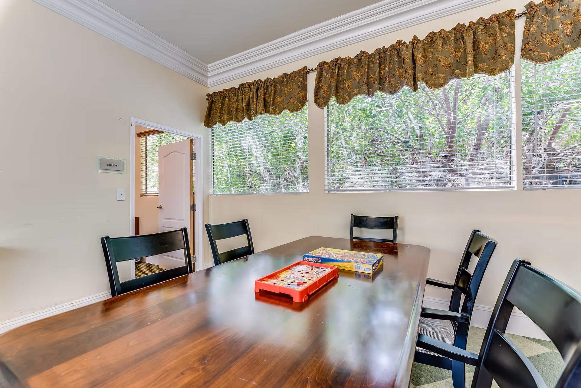 A bright room with a wooden table surrounded by six black chairs. On the table, there are two board games. Large windows with blinds and floral valances let in natural light and show green trees outside. A door labeled 'Library' is open, revealing another room.