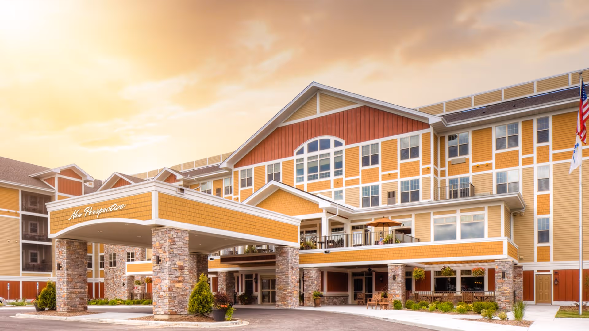 Exterior view of New Perspective Senior Living facility in Sun Prairie during sunset, showing a multi-story building with yellow and red siding, stone pillars, a covered entrance, and an American flag on the right side.