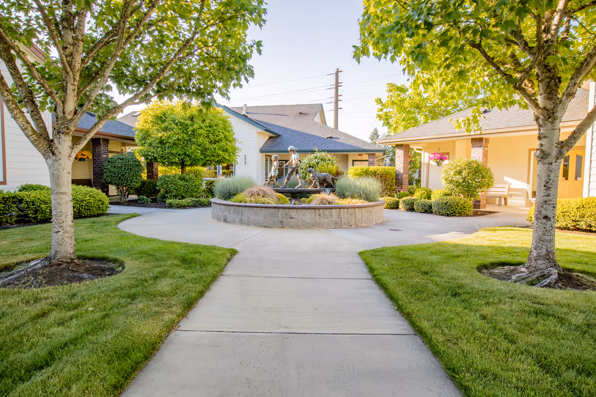 A sunny courtyard at Gateway Gardens featuring a circular stone planter with a bronze statue of children and a dog in the center. The courtyard is surrounded by well-maintained green lawns, trees, shrubs, and single-story buildings with covered walkways and benches.
