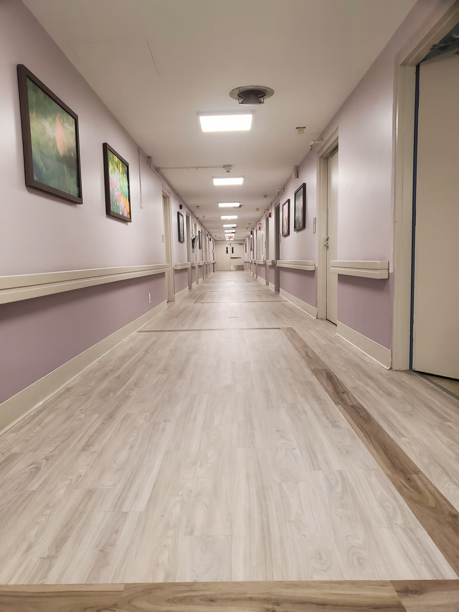 Long indoor hallway with light wood flooring and lavender walls, decorated with framed pictures on both sides. The hallway has multiple doors and ceiling lights evenly spaced along the length.