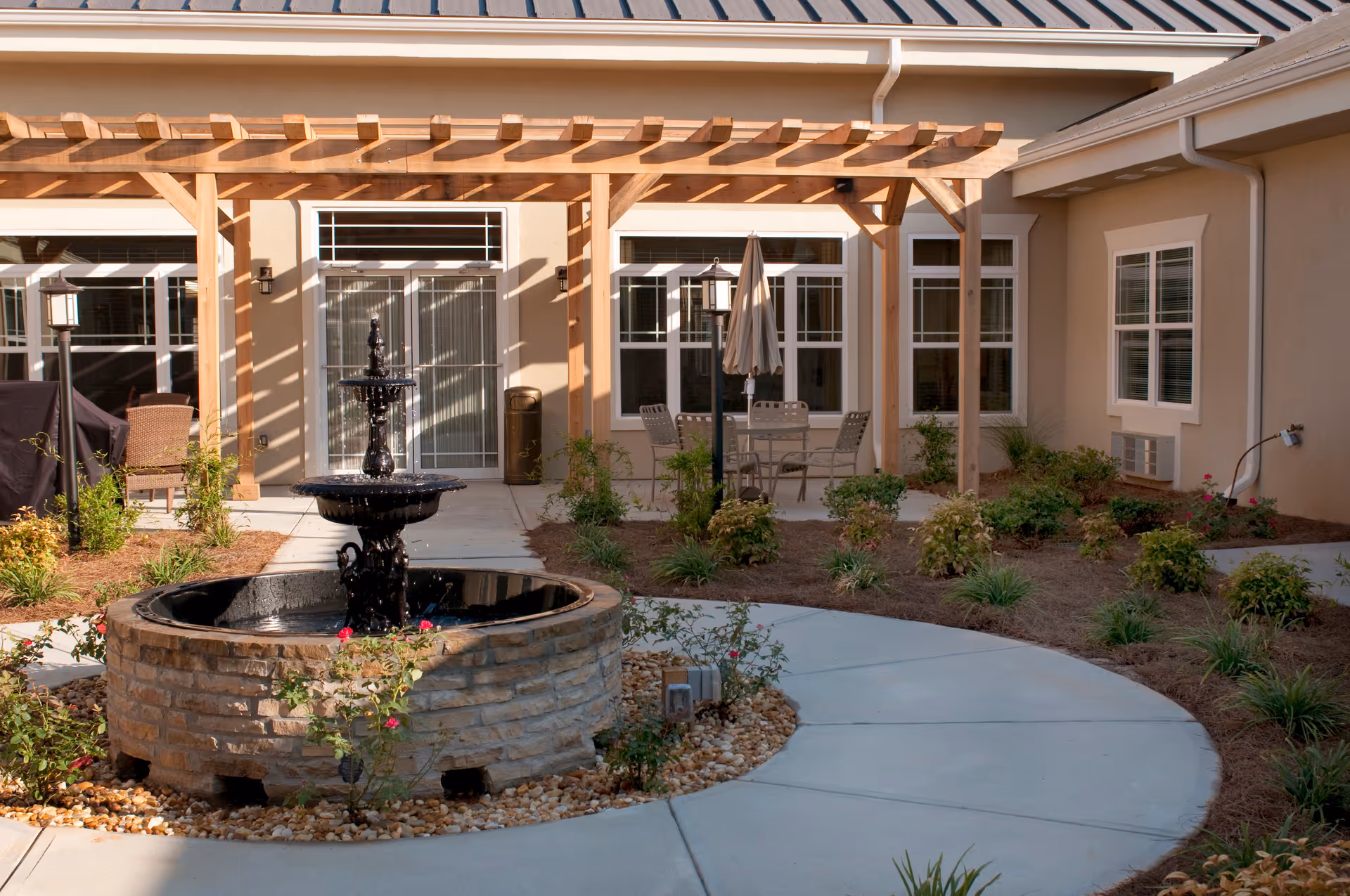 Outdoor courtyard area with a stone water fountain in the center, surrounded by a circular concrete walkway and landscaped plants. There is a wooden pergola structure attached to the building, with patio furniture including a table, chairs, and an umbrella. The building has beige walls and multiple windows.