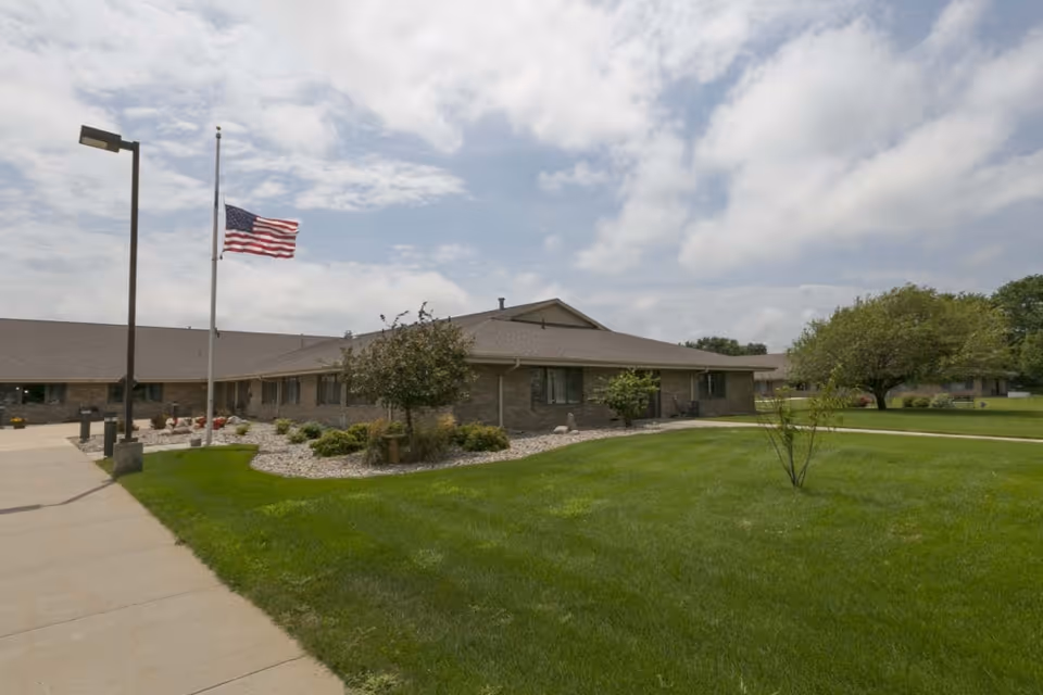 Exterior view of a single-story brick building with a well-maintained lawn and landscaping. An American flag is flying at half-staff on a flagpole near the sidewalk. The sky is partly cloudy.