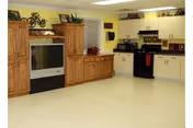 Bright common room in a nursing center with a large TV cabinet, storage cabinets, and a small kitchenette with a stove and microwave.