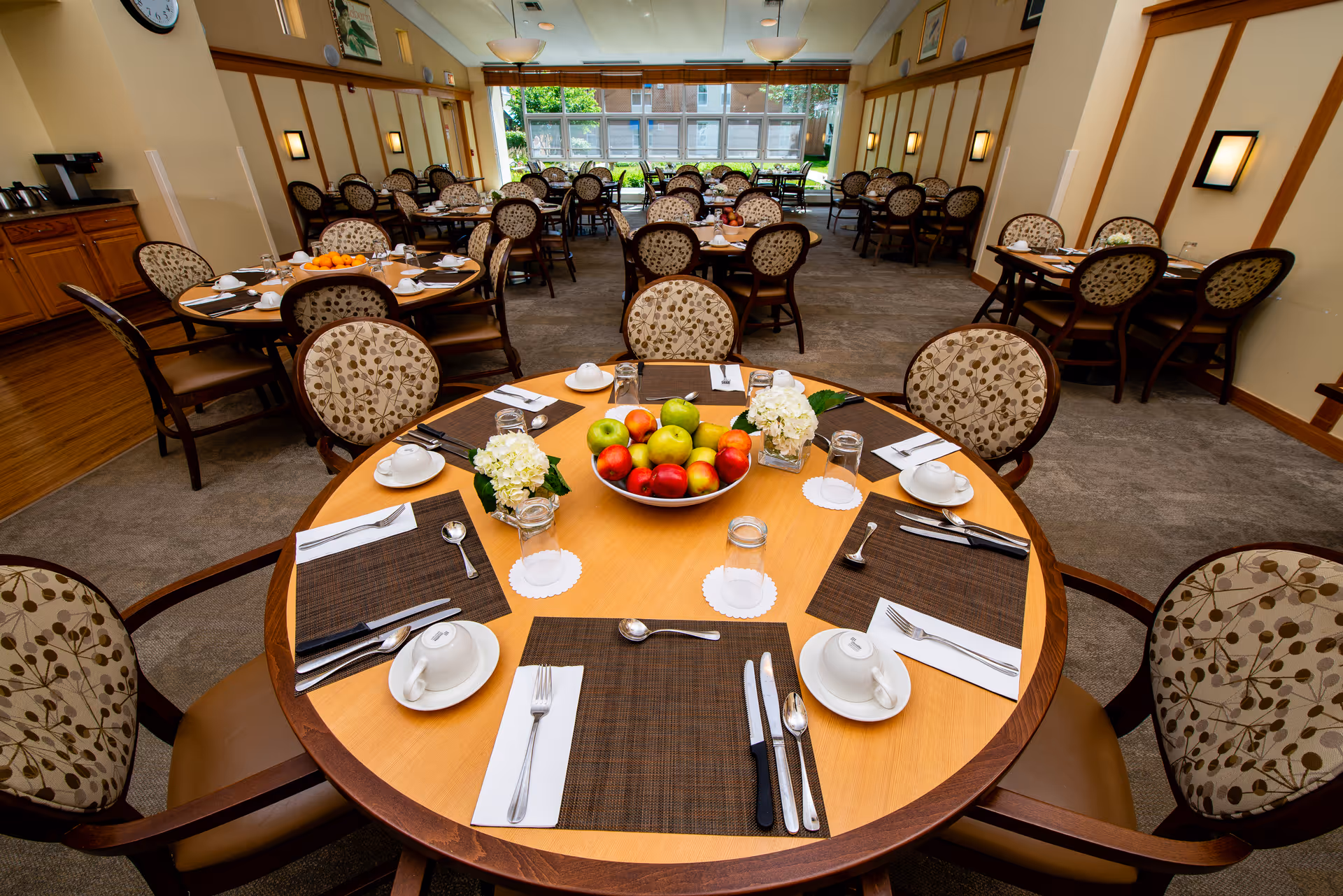 A well-lit dining room with multiple round tables set for meals. Each table has placemats, utensils, cups, and napkins arranged neatly. The table in the foreground has a centerpiece bowl filled with apples and two small flower arrangements. The room has patterned chairs, wall sconces, large windows letting in natural light, and a clock on the wall.