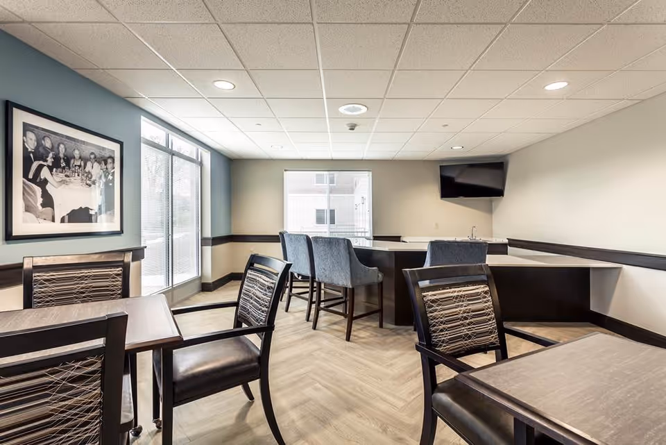A modern common area with several tables and chairs, a counter with high chairs, a wall-mounted TV, large windows letting in natural light, and a framed black-and-white photo on the wall.