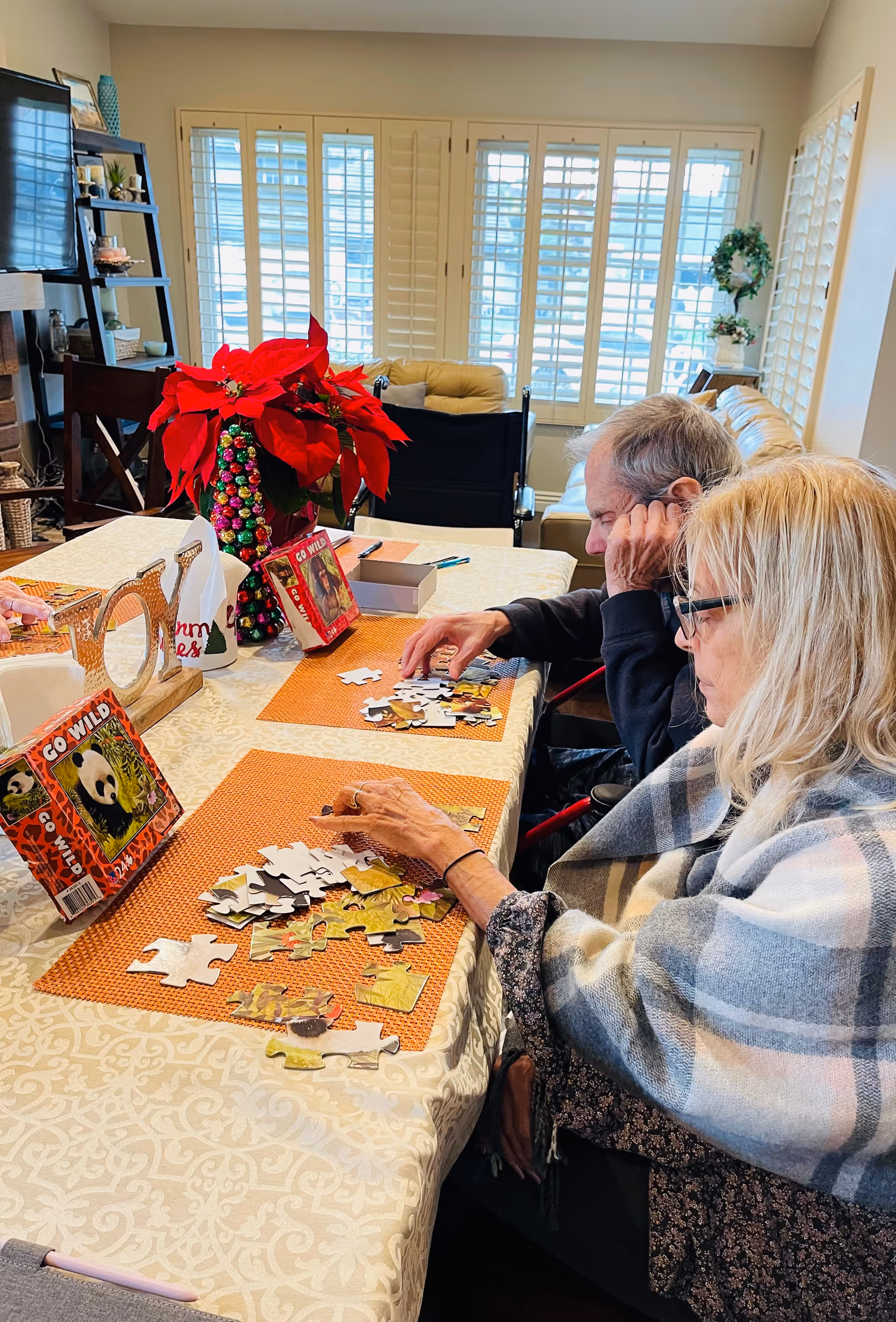 Two elderly people sitting at a dining table working on jigsaw puzzles with a red poinsettia centerpiece in a bright room.