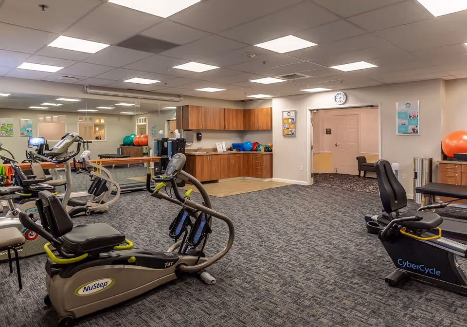 A senior fitness room with exercise equipment including NuStep and CyberCycle machines, a large mirror on one wall, wooden cabinets with exercise balls on the countertop, and a doorway leading to another room. The room has carpeted flooring and a drop ceiling with fluorescent lights.