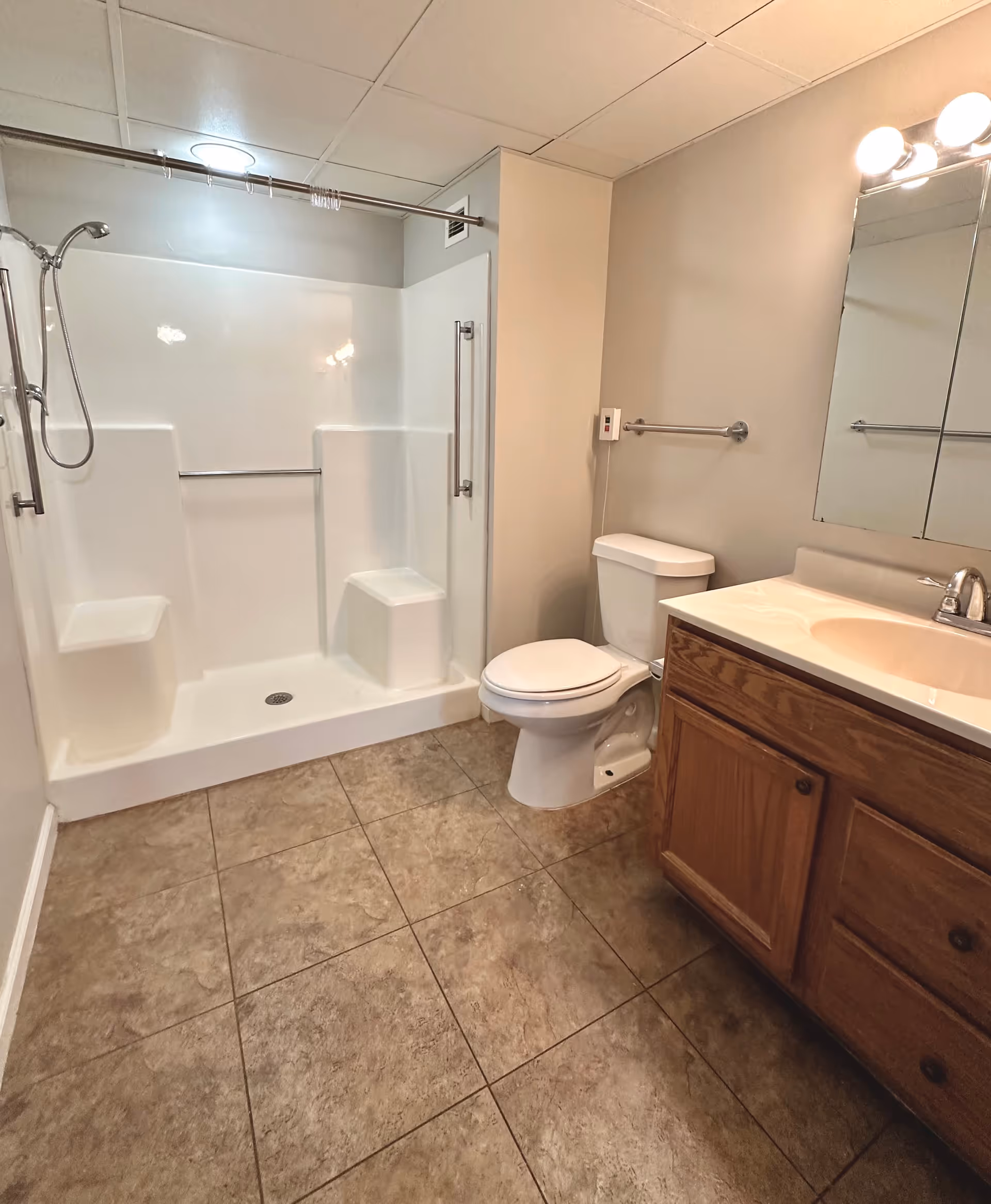 A clean bathroom with a walk-in shower featuring built-in seats and grab bars, a toilet, a wooden vanity with a sink, and a large mirror above the sink. The floor is tiled and the walls are painted a light beige color.