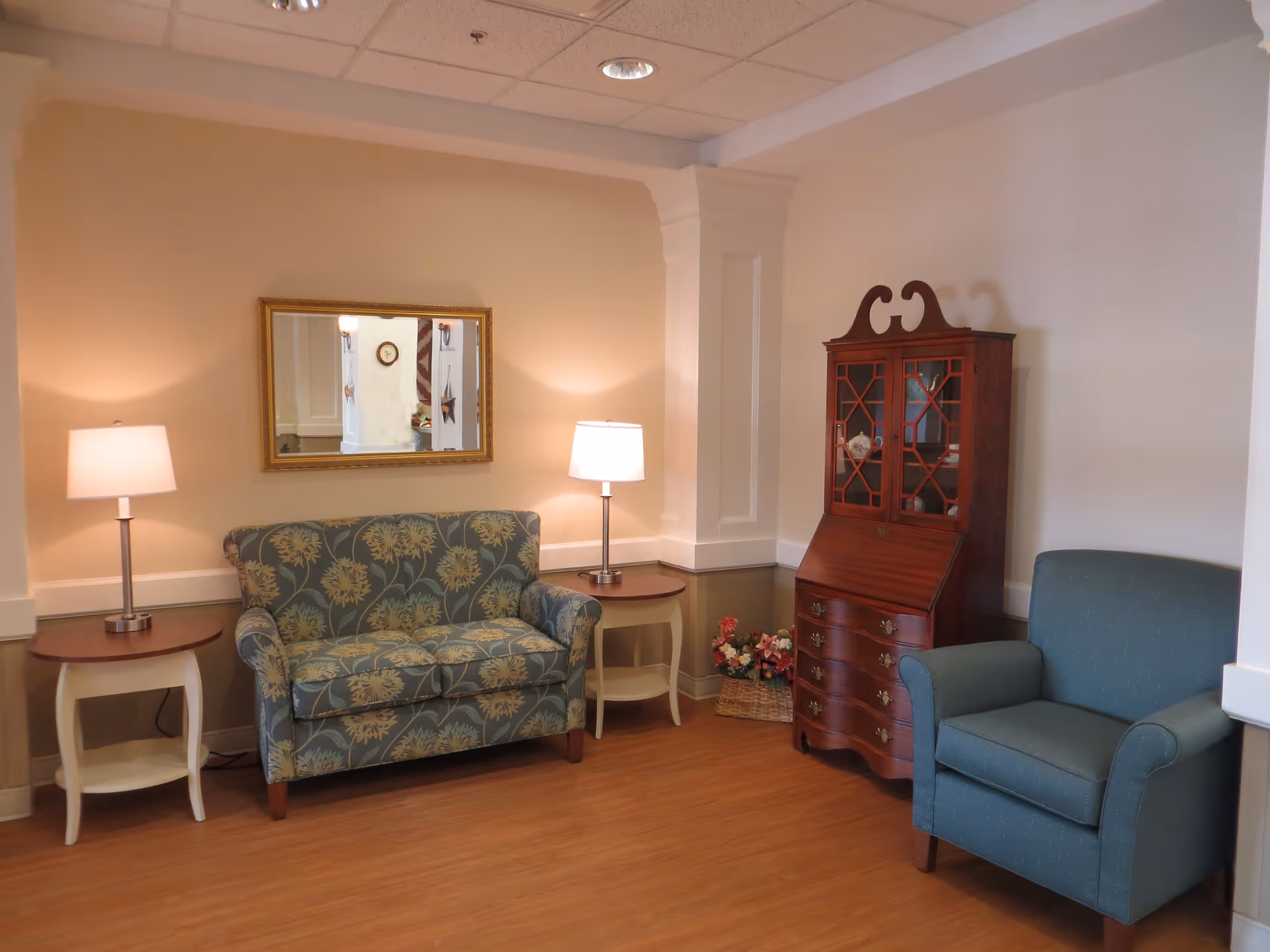 Cozy seating area with a patterned loveseat, armchair, side tables with lamps, and a wooden cabinet against a beige wall.