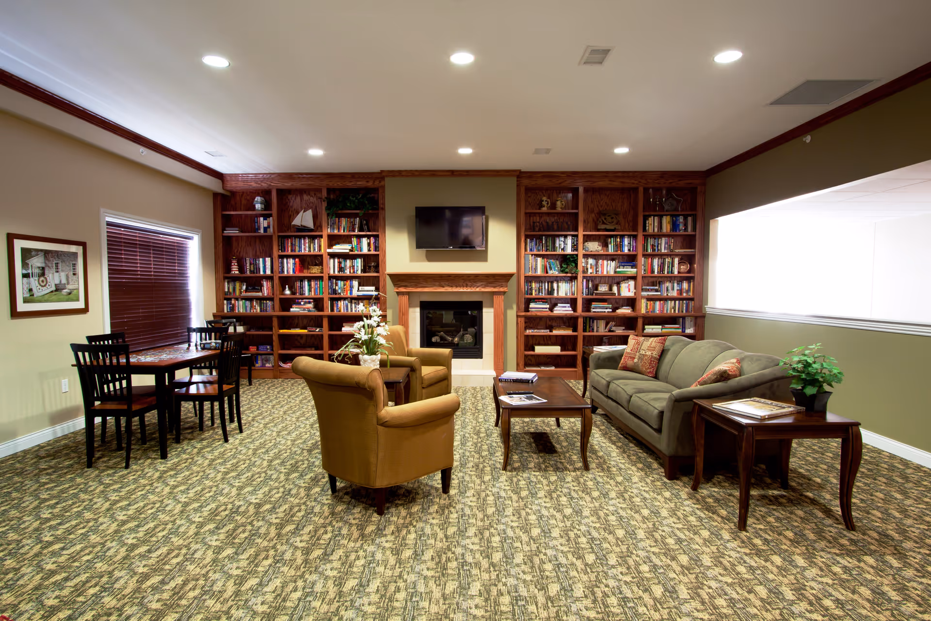 A cozy living room area in a retirement community featuring a green sofa with patterned pillows, two brown armchairs, a wooden coffee table, and side table with a potted plant. Behind the seating area is a fireplace with a mounted flat-screen TV above it, flanked by large wooden bookshelves filled with books and decorative items. To the left, there is a small dining table with four chairs near a window with closed blinds. The room has patterned carpet flooring and beige walls with white trim.