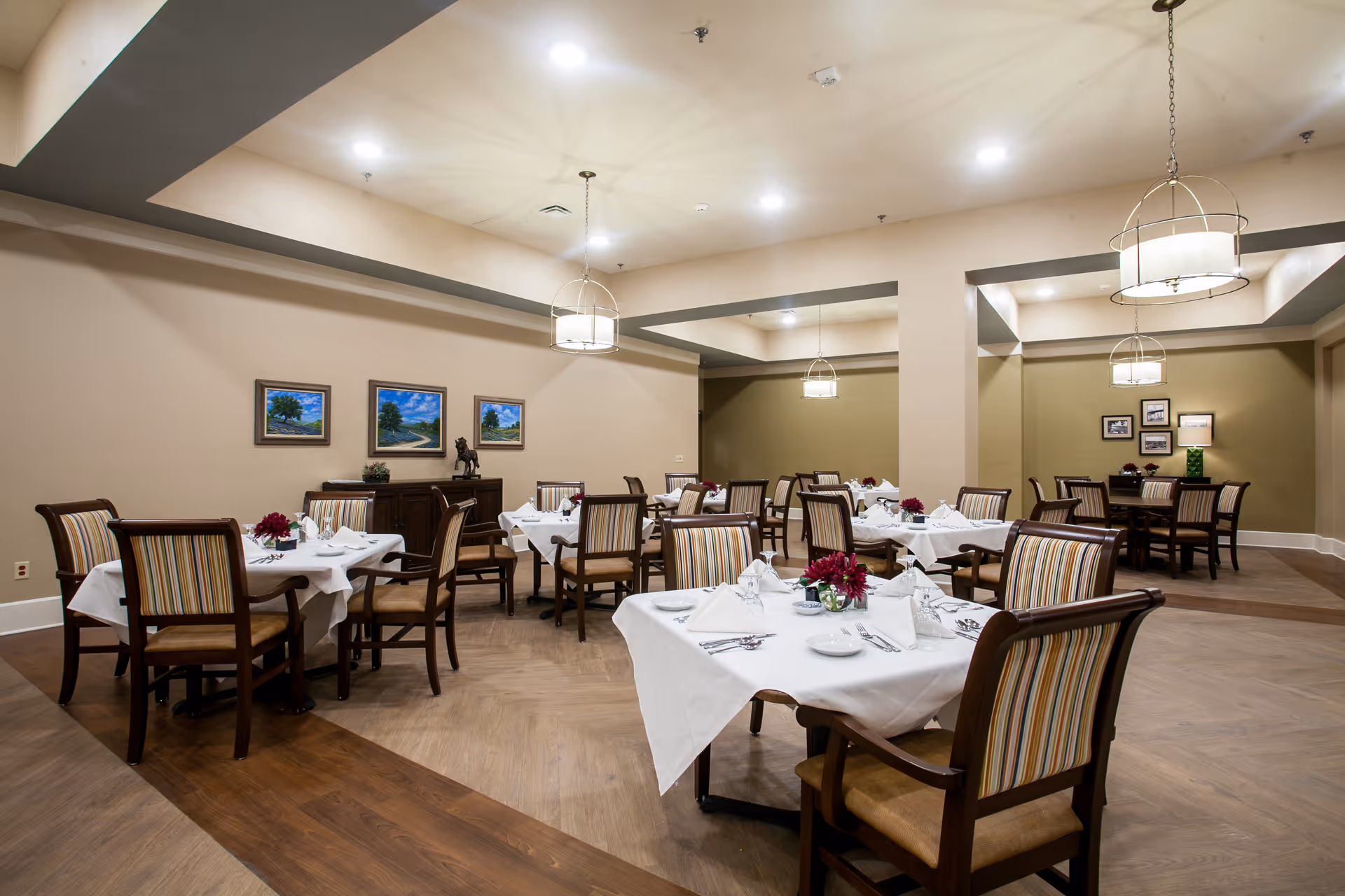 A well-lit dining room with multiple tables covered in white tablecloths, each set with napkins, plates, and silverware. The chairs have striped upholstery, and there are small floral centerpieces on each table. The walls are decorated with framed landscape paintings and photographs. Modern pendant lights hang from the ceiling, illuminating the space.