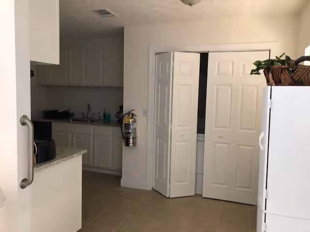 Interior view of a kitchen area with white cabinets, a countertop with a microwave, a fire extinguisher mounted on the wall, and bi-fold doors partially open revealing a laundry area. A white refrigerator with a plant on top is visible on the right side.