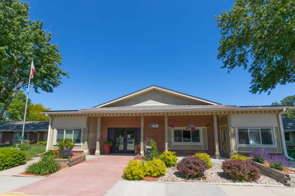 Front exterior view of Fort Dodge Health and Rehabilitation building with a clear blue sky, landscaped bushes and flowers, an American flag on a pole to the left, and a paved walkway leading to double glass entrance doors.