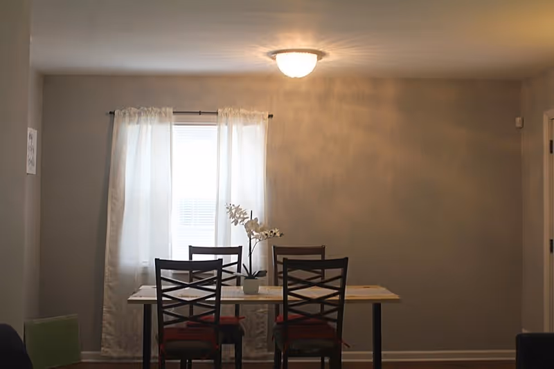 A dining area with a rectangular table and four chairs with red cushions. A small potted orchid is placed in the center of the table. Behind the table is a window with white curtains, and a ceiling light fixture is turned on above the table. The walls are painted light gray.