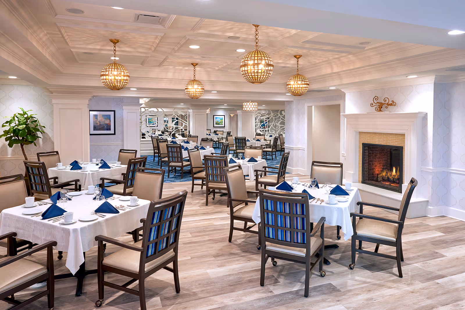 A spacious, elegant dining room with multiple tables covered in white tablecloths, each set with blue napkins, white cups, and silverware. The room features wooden chairs with blue and beige upholstery, a lit fireplace on the right wall, decorative hanging light fixtures, and a large plant in the corner. The walls are light-colored with subtle patterns, and mirrors are visible in the background.