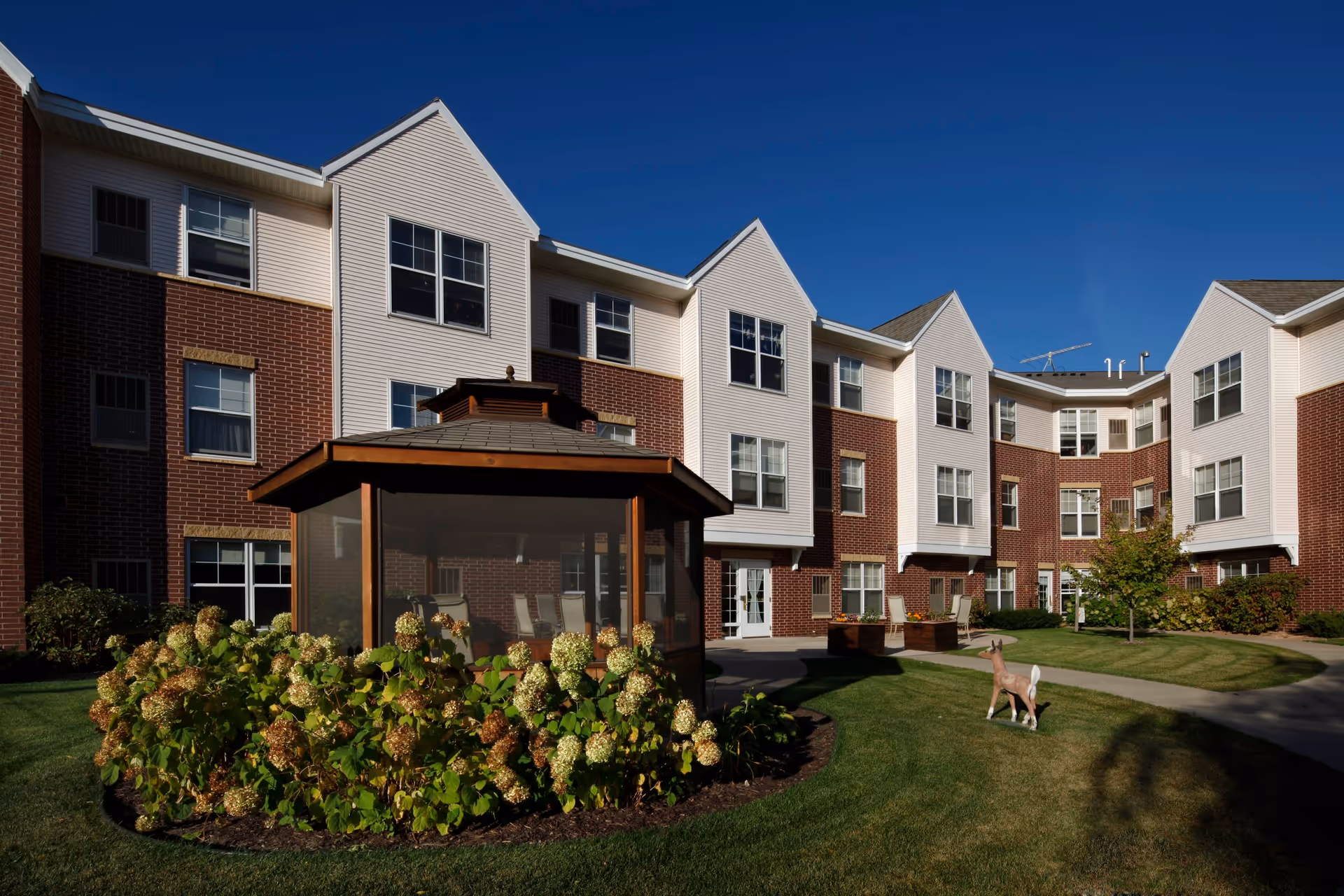 Exterior view of a senior living facility building with three stories, featuring a combination of brick and white siding. In front of the building, there is a landscaped garden with green grass, flowering bushes, a small tree, and a wooden gazebo with seating inside. A small decorative deer statue is also visible on the lawn under a clear blue sky.
