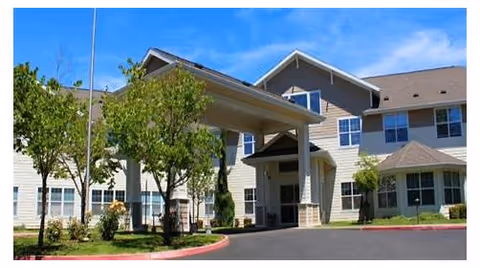 Exterior view of a multi-story senior living facility building with beige siding and a covered entrance. There are several windows, small trees, and shrubs around the building, with a clear blue sky in the background.