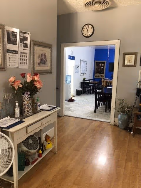 Interior view of a senior living facility showing a hallway with wooden flooring leading to a dining area with tables and chairs. On the left side of the hallway is a small white table with a vase of pink and red flowers, hand sanitizer, and some papers. The walls are decorated with framed pictures and notices. A clock is mounted above the doorway to the dining area, which has tiled flooring and blue accent walls.
