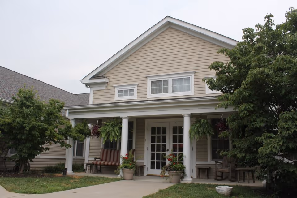 Front entrance of a beige two-story assisted living building with white columns, a covered porch, hanging ferns, and potted plants.