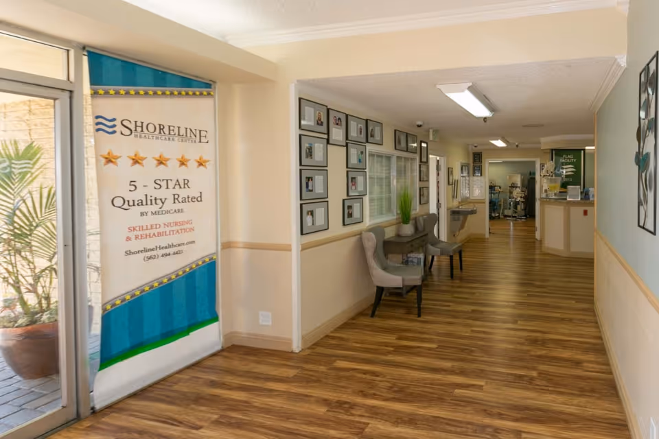 Interior hallway of Shoreline Healthcare Center with wood flooring, beige walls, and framed photos on the wall. Two upholstered chairs and a small table with a plant are placed along the hallway. A banner near the entrance highlights the facility's 5-star quality rating by Medicare for skilled nursing and rehabilitation.