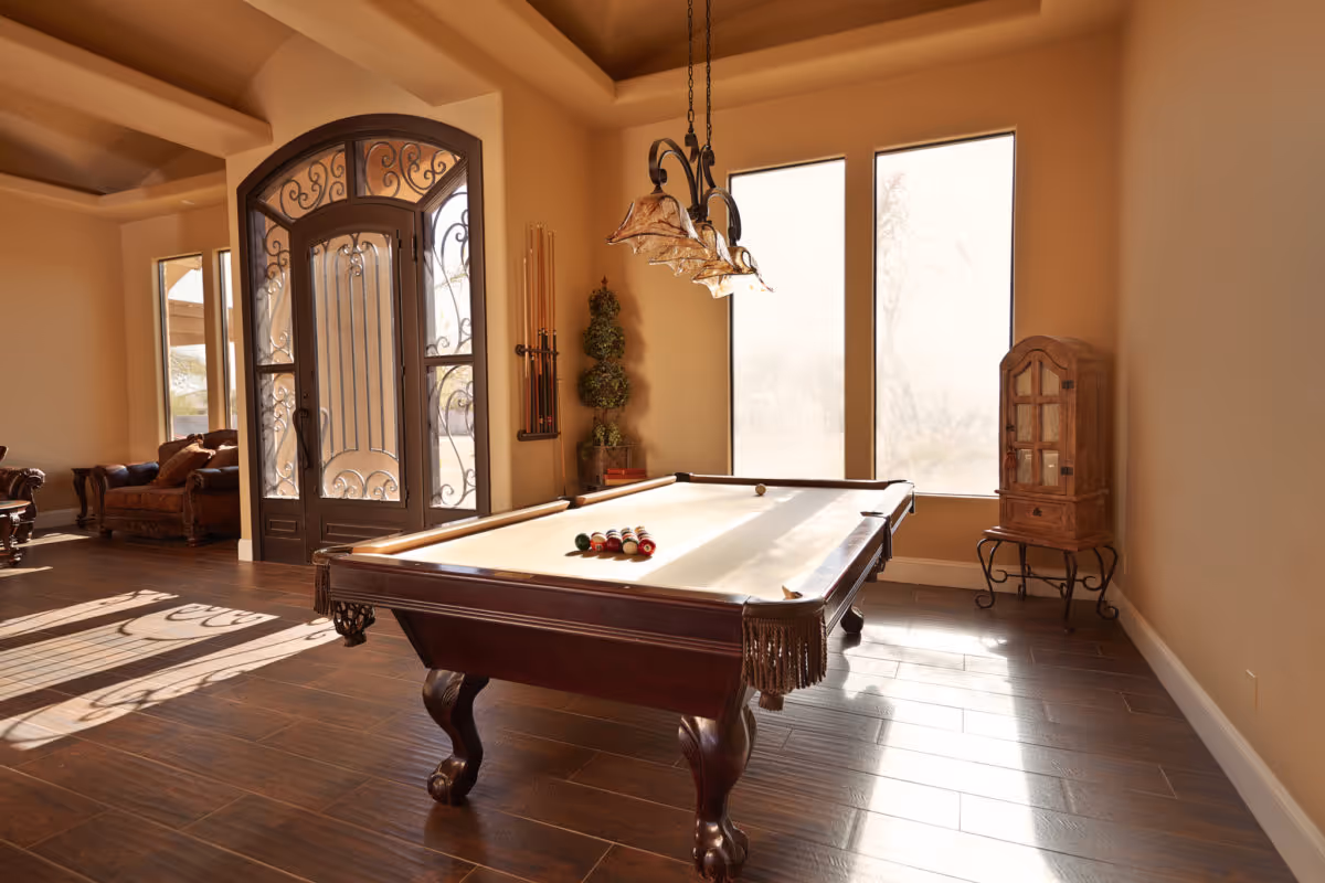 A well-lit room with a pool table in the center, a set of billiard balls arranged on the table, large windows letting in natural light, a decorative wooden cabinet on the right, and an ornate glass door with wrought iron details on the left. The room has wooden flooring and warm beige walls.