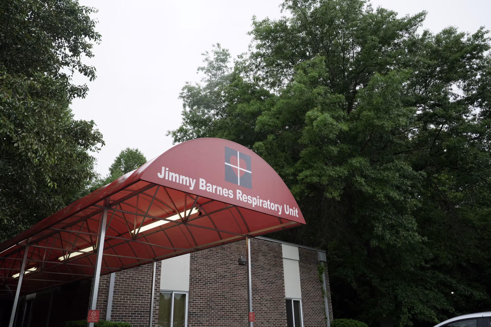 Entrance canopy with a red roof labeled 'Jimmy Barnes Respiratory Unit' attached to a brick building surrounded by green trees.