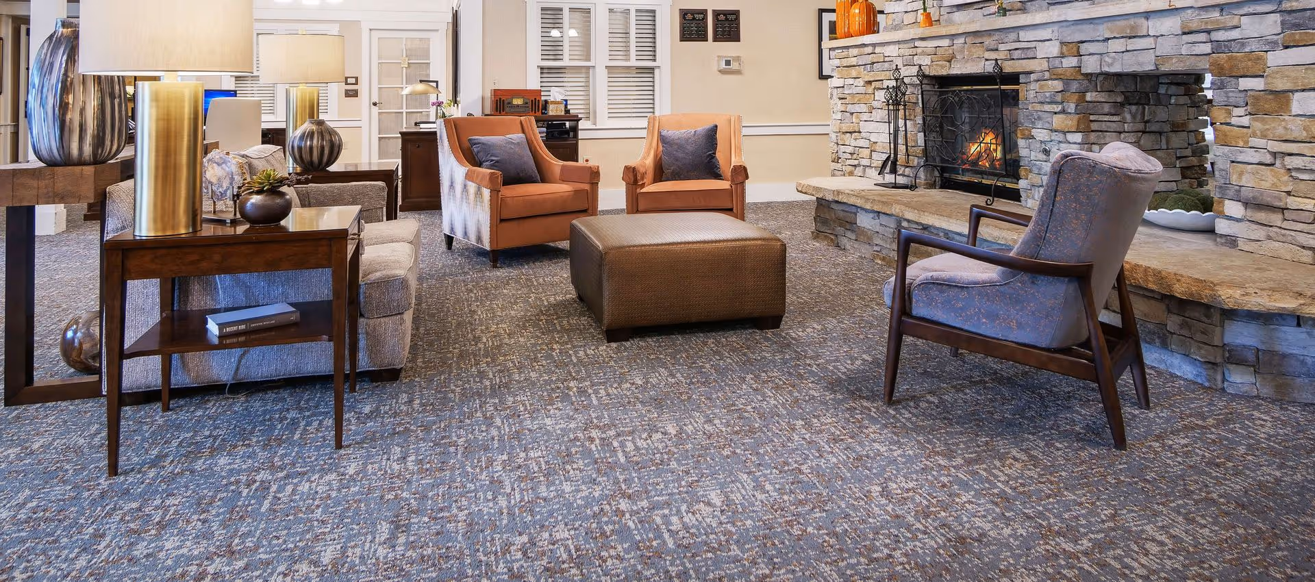 A cozy living room area with a stone fireplace on the right side, a fire burning inside. The room has a patterned carpet floor, two orange armchairs with blue cushions, a gray armchair with wooden arms, and a large brown ottoman in the center. There are wooden side tables with lamps and decorative items, and a window with white shutters in the background.