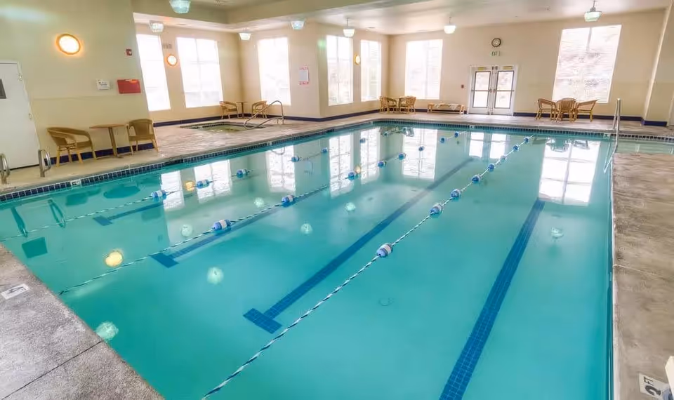Bright indoor lap pool with lane dividers in a senior living facility activity room with chairs and large windows.