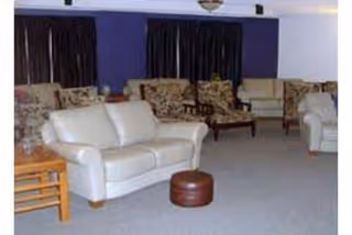 A living room area with multiple upholstered chairs and sofas arranged on a carpeted floor. The walls are painted purple and white, and there are dark curtains covering the windows.