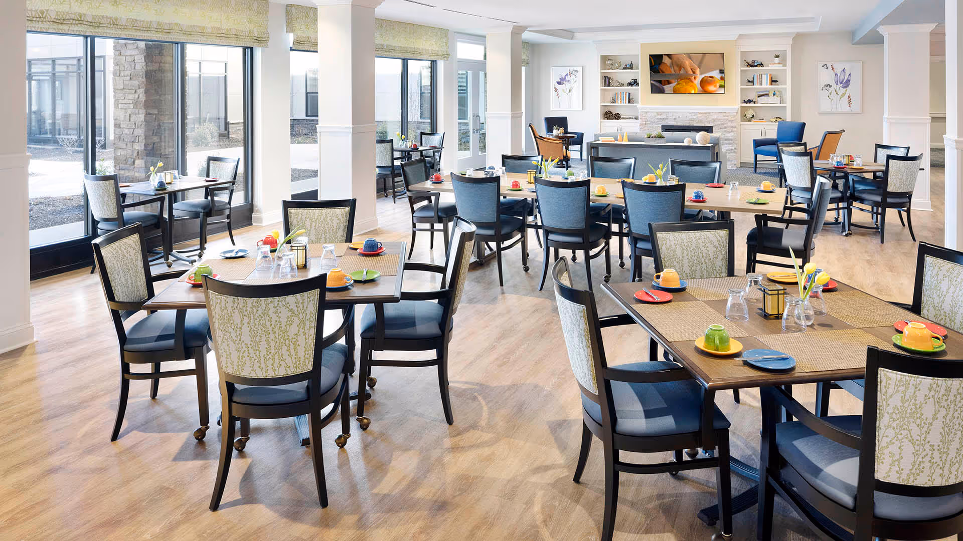 Bright dining room with multiple tables and chairs arranged neatly. Each table has colorful cups and plates, placemats, and small flower vases. Large windows on the left side let in natural light. A fireplace with built-in shelves and framed artwork is visible on the far wall.