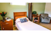 A single-occupancy bedroom with a bed, wooden headboard, nightstand, desk with chair, and an armchair against light green walls.
