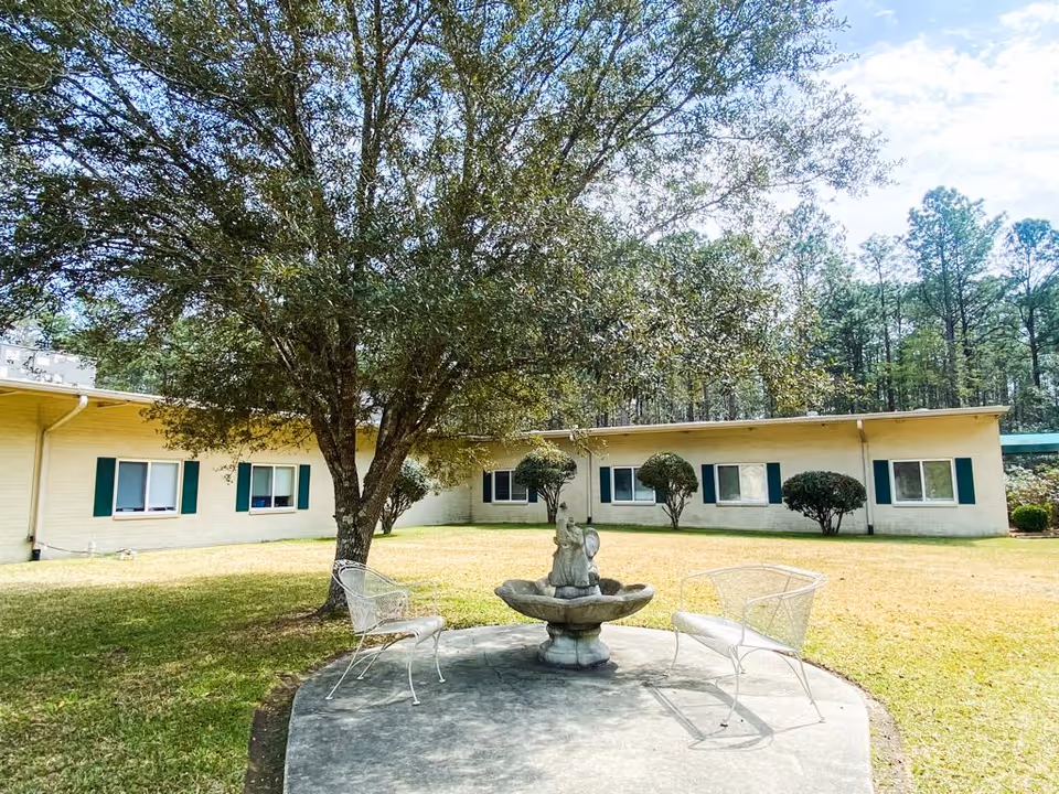 Outdoor courtyard area at Picayune Rehabilitation and Healthcare Center featuring a large tree, a small decorative fountain, and two white metal chairs on a concrete patio. The building with multiple windows and green shutters surrounds the grassy area, with trees visible in the background under a partly cloudy sky.