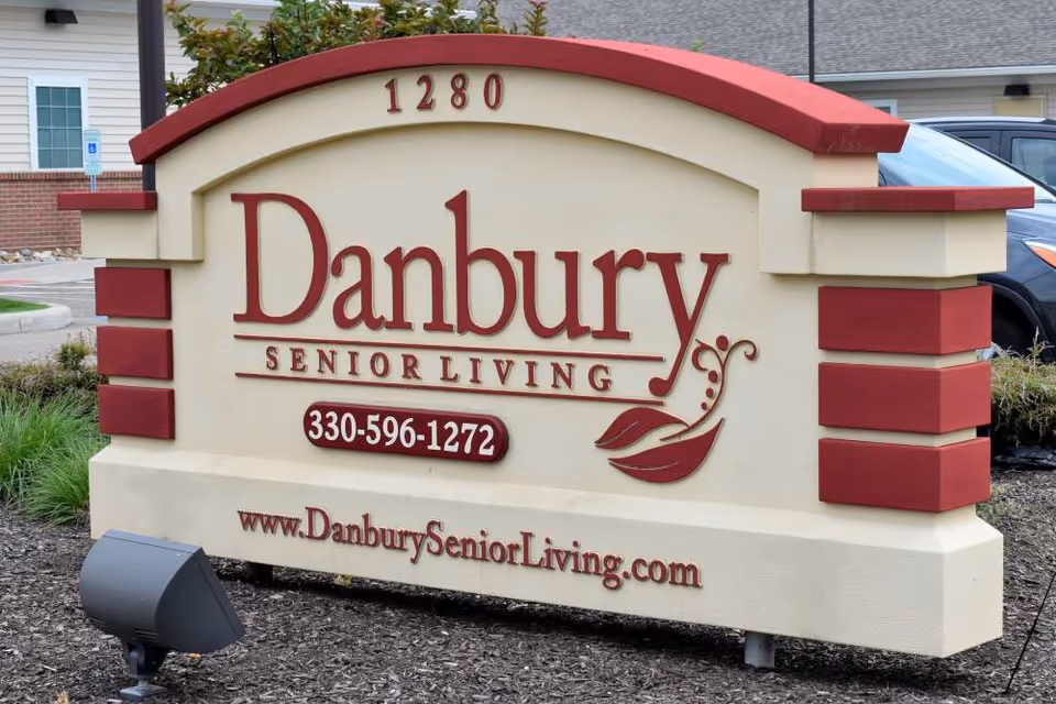 Outdoor sign for Danbury Senior Living facility with the address number 1280, phone number 330-596-1272, and website www.DanburySeniorLiving.com. The sign is cream-colored with red accents and decorative elements, situated in a landscaped area with mulch and plants.